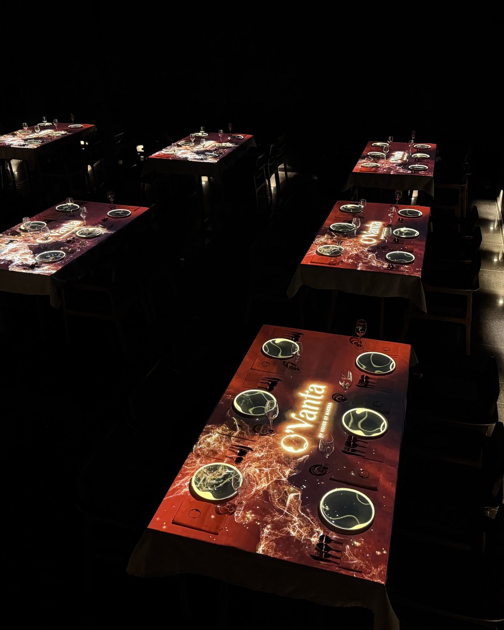 The O'Vanta dining room: rows of projection-mapped tables glowing in the dark, the O'Vanta mark cast across the nearest table